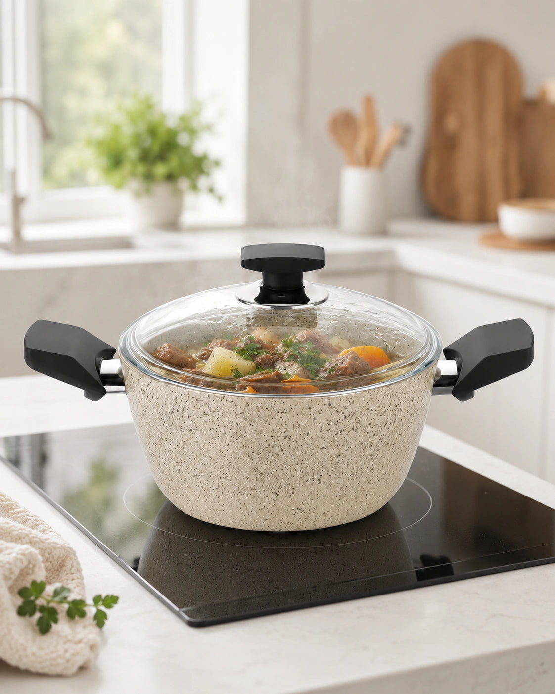 A speckled beige non-stick pot with a clear glass lid and black handles sits on a black induction cooktop in a bright, modern kitchen.