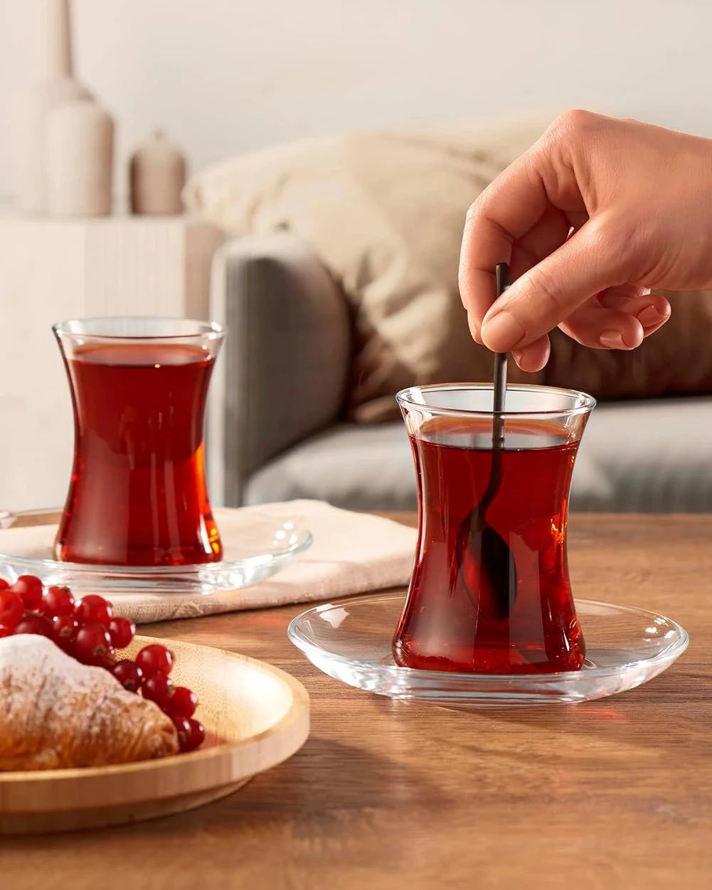 Person stirring tea in a glass with a spoon on a wooden table, with a plate of grapes and a pastry in the foreground.